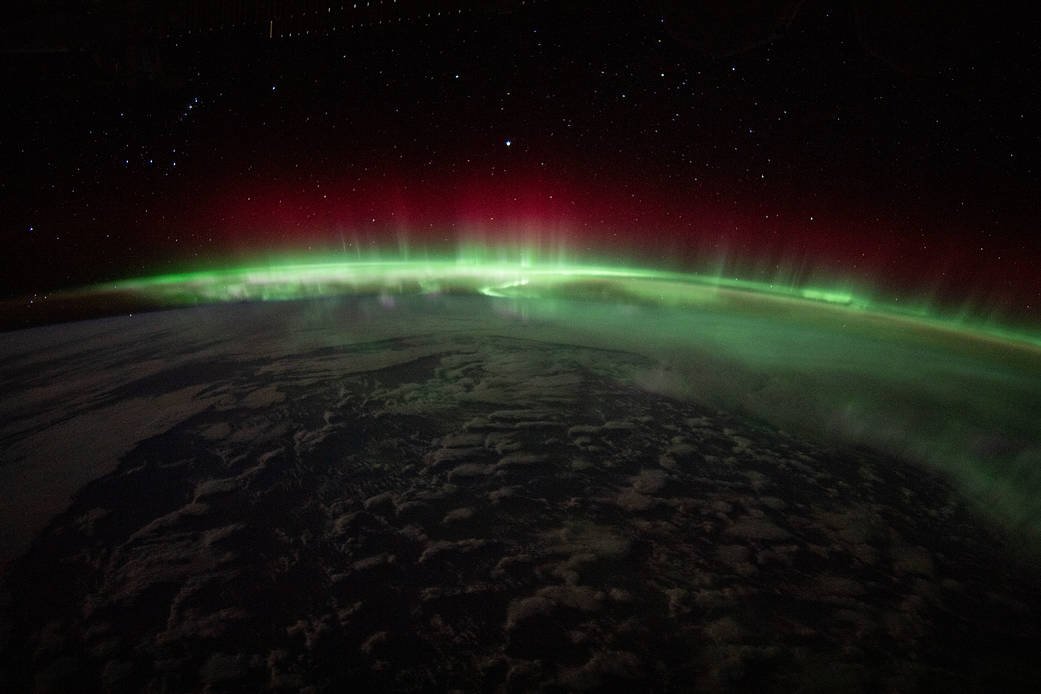 A green and red aurora glows along Earth’s curved horizon beneath a starry sky.