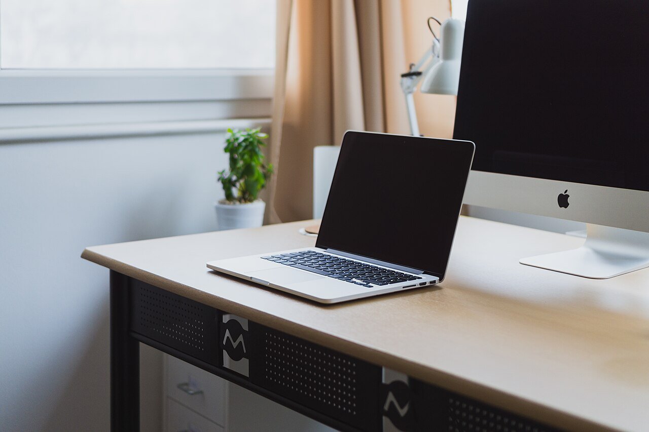 An open silver laptop sits on a tidy white desk beside a desktop monitor.