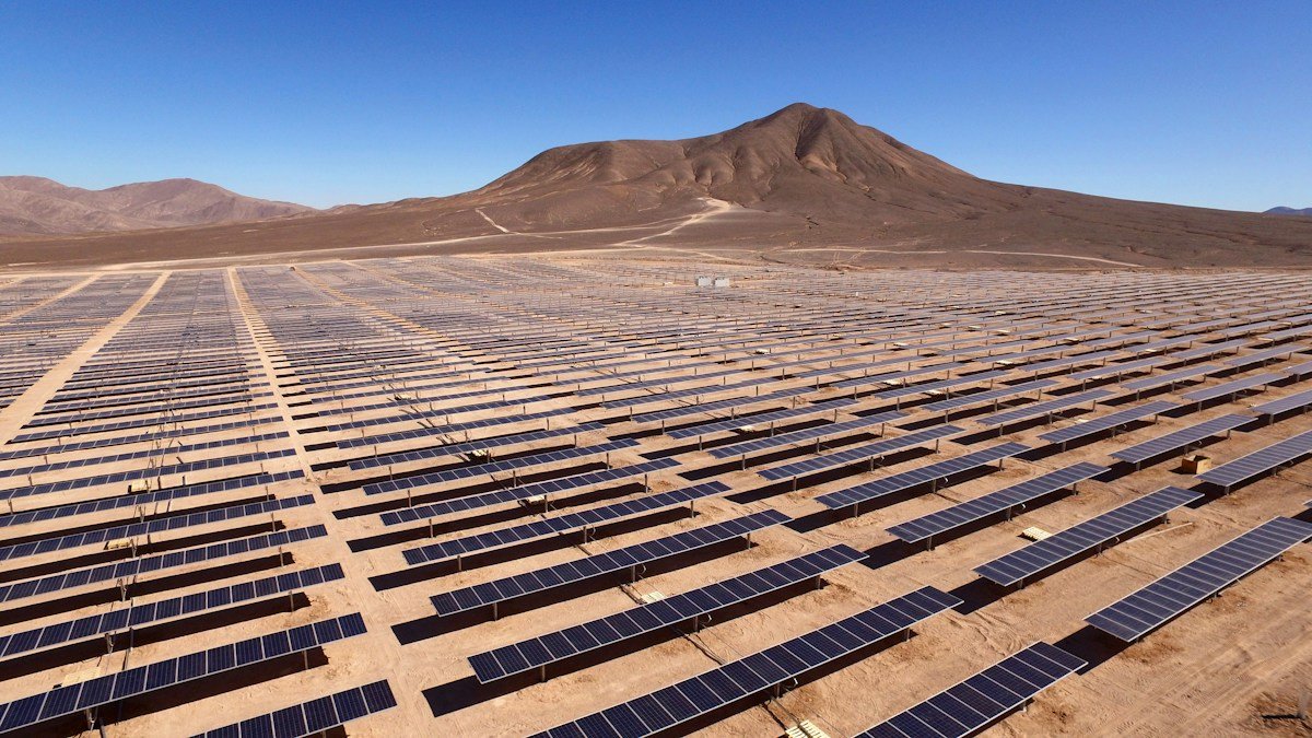 Long rows of solar panels cover a desert site beneath a mountain and clear blue sky.